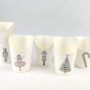 May include: Off-white paper cups with a curved top, featuring illustrations of a nutcracker, ballerina, mouse, Christmas tree, and candy cane. The cups are stacked and arranged on a white surface, ideal for holiday gatherings.