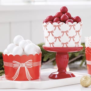 May include: Red and white paper cupcake liners with a bow pattern. The liners are filled with white and red candies. The liners are on a red pedestal and a white surface.