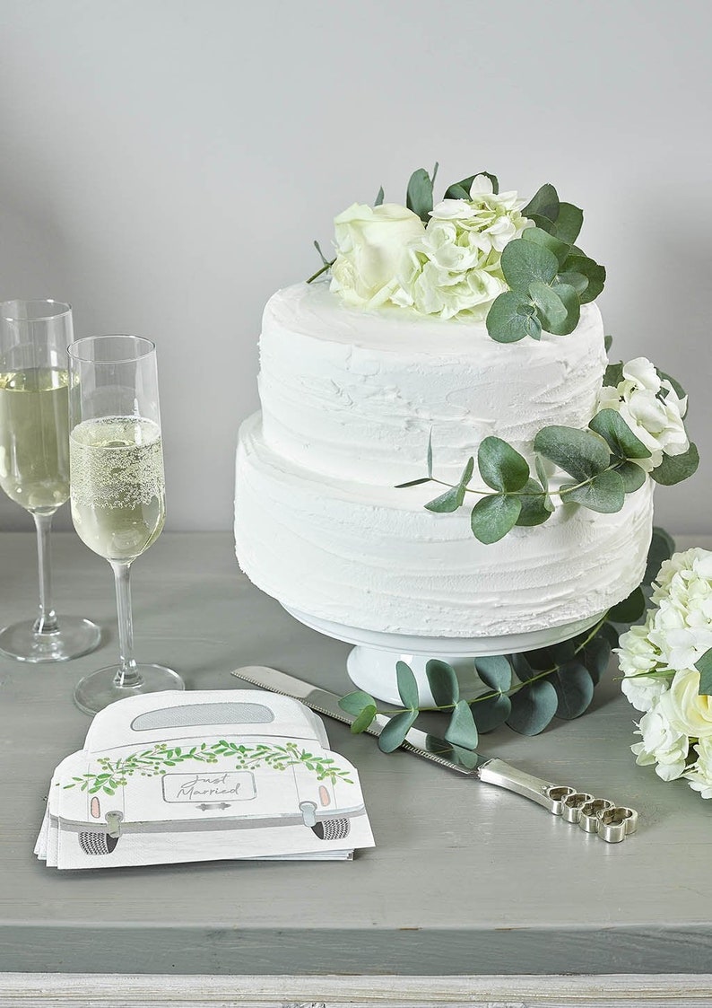 May include: A two-tiered white wedding cake decorated with white flowers and greenery. The cake is on a white cake stand. There are two champagne flutes and a cake knife on the table. There are also napkins with a car design and the text "Just Married" on them.