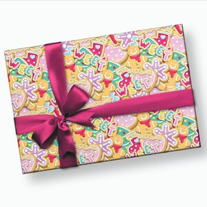 May include: A pink and white gift box with a pink ribbon and bow. The box is wrapped in paper with a pattern of gingerbread cookies with the words "Sweet" and "Christmas" on them.