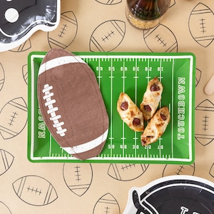May include: A football-themed party setting with a green field-shaped tray, a football-shaped napkin, and snacks. The background features a football pattern. Black and white plates with the words "GO!" and "LET GO" are also visible.