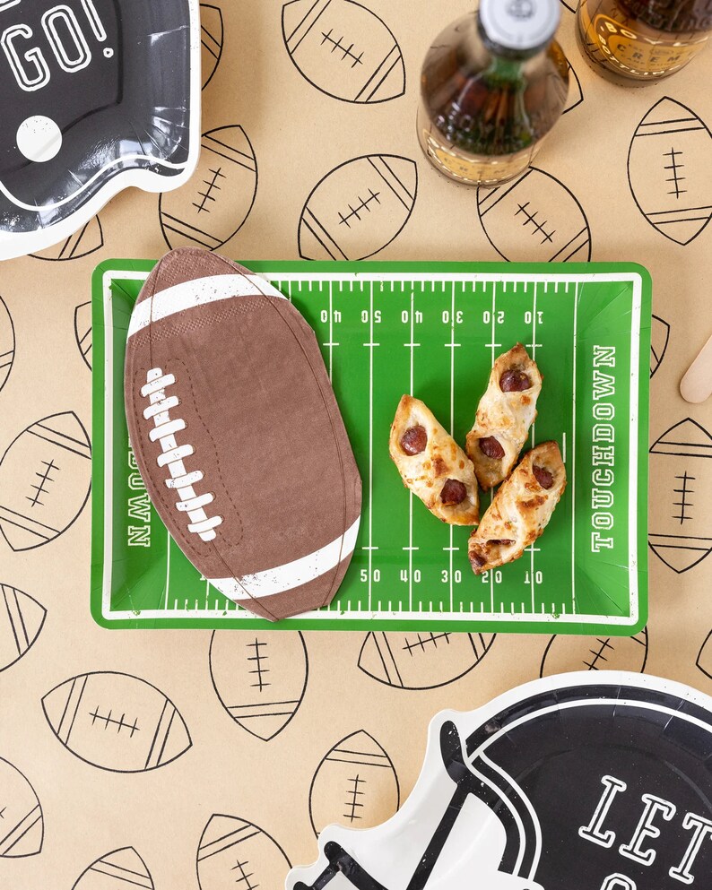 May include: A football-themed party setting with a green field-shaped tray, a football-shaped napkin, and snacks. The background features a football pattern. Black and white plates with the words "GO!" and "LET GO" are also visible.