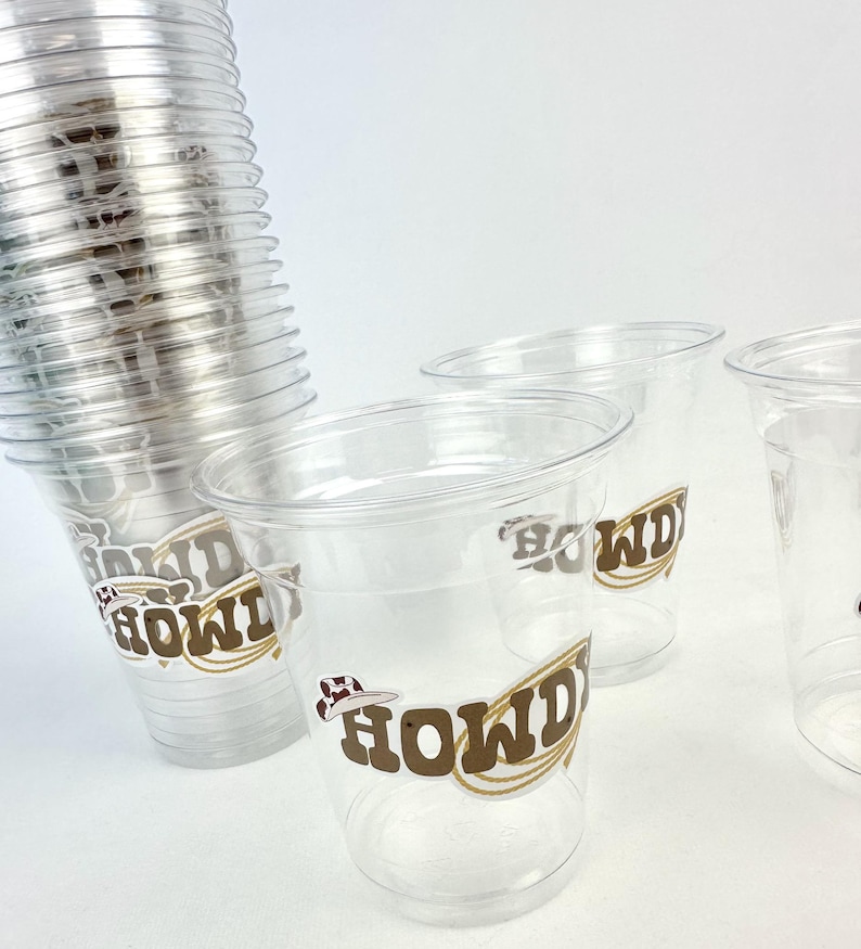 Puede incluir: Vasos de pl&aacute;stico transparente con un gr&aacute;fico "HOWDY" en marr&oacute;n y blanco. Los vasos tienen un dise&ntilde;o de sombrero de vaquero. Una pila de vasos es visible a la izquierda, con m&aacute;s vasos en primer plano.