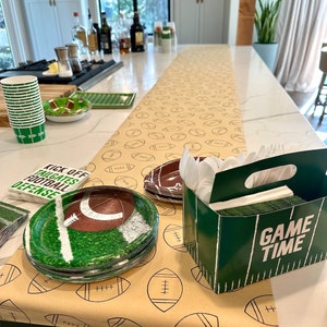 May include: A football-themed party setup with a tan table runner printed with footballs. Includes green and white paper cups, plates with football and field designs, napkins, and a green box that says "GAME TIME".