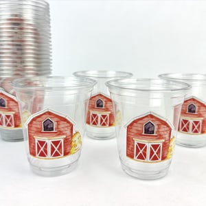 May include: Clear plastic cups with a red barn design. The barn has white doors and a purple window. A stack of cups is in the background. The cups are suitable for a farm-themed party or event.