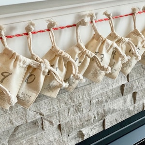 Puede incluir: Calendario de Adviento con peque&ntilde;as bolsas beige con cord&oacute;n, numeradas del 1 al 16, colgadas de una cuerda roja y blanca. Cada bolsa tiene un dise&ntilde;o de copo de nieve y las palabras "Enjoy the Season". Las bolsas est&aacute;n contra una pared de piedra.