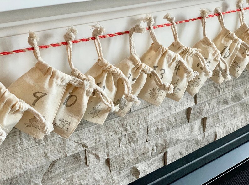 Puede incluir: Calendario de Adviento con peque&ntilde;as bolsas beige con cord&oacute;n, numeradas del 1 al 16, colgadas de una cuerda roja y blanca. Cada bolsa tiene un dise&ntilde;o de copo de nieve y las palabras "Enjoy the Season". Las bolsas est&aacute;n contra una pared de piedra.