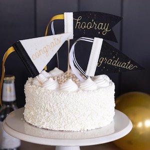 May include: A white cake with white frosting and sprinkles, topped with three decorative pennant flags. The flags are black and white with gold lettering that reads "congrats", "hooray", and "graduate". The cake sits on a white cake stand.