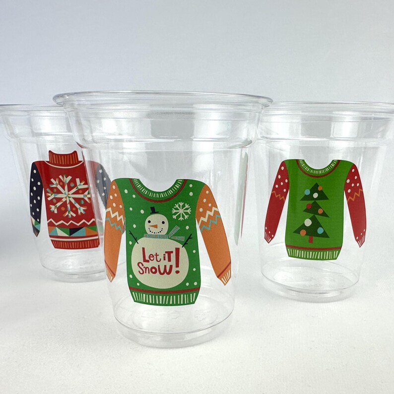 May include: Clear plastic cups with festive Christmas sweater designs. One cup features a red sweater with a snowflake, another a green sweater with a snowman and the text "Let it Snow!", and a third with a green sweater and Christmas trees.