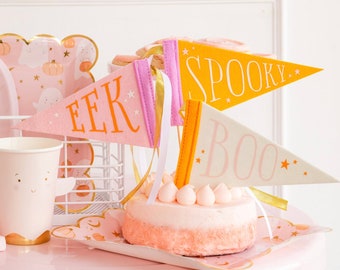 Puede incluir: Un pastel rosa y blanco con banderas naranja y amarillas que dicen "EEK", "BOO" y "SPOOKY" en la parte superior. El pastel está en un plato rosa y blanco.