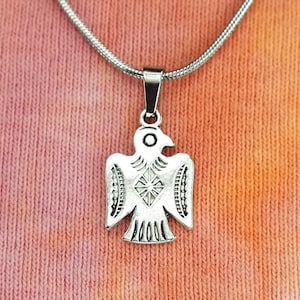 May include: A silver pendant necklace with a bird motif. The pendant, suspended from a fine silver chain, depicts a stylized bird with detailed wings and body. The background is a tie-dye pattern in shades of orange and pink.