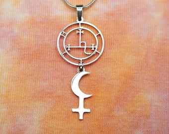 Puede incluir: Un collar de plata con un colgante que presenta un círculo con un símbolo en su interior y una luna creciente colgando de él. El collar está sobre un fondo naranja y rosa tie-dye.