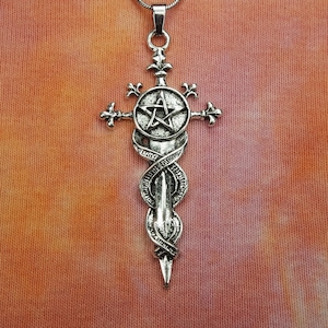 May include: A silver-toned pendant necklace featuring a cross design with a pentagram at the center. The pendant has intricate details, including a sword-like element and decorative flourishes. The necklace hangs on a thin silver chain.