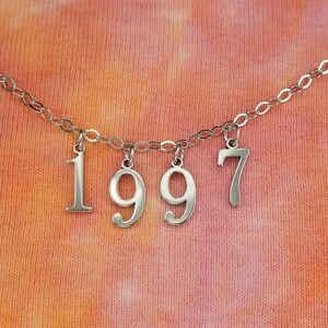 以下が含まれることがあります： 銀色のチェーンネックレスに、1997の数字の形をした銀色のチャームが付いています。ネックレスは、薄いオレンジ色とピンク色のタイダイ模様の背景の上に置かれています。