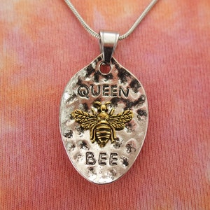May include: Silver-toned pendant necklace with a hammered texture. The pendant features the words "QUEEN BEE" and a gold-colored bee design. The necklace has a silver chain and a lobster clasp.