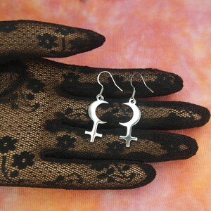May include: Silver-toned earrings featuring a crescent moon design above a Venus symbol. The earrings are displayed against a backdrop of a black lace glove and a pink and orange gradient background.