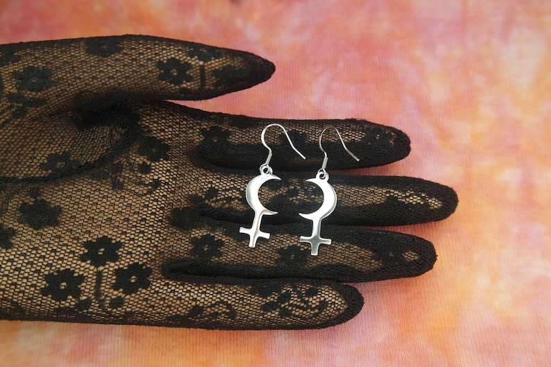 May include: Silver-toned earrings featuring a crescent moon design above a Venus symbol. The earrings are displayed against a backdrop of a black lace glove and a pink and orange gradient background.