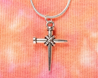 May include: A silver necklace with a cross pendant. The cross is made to look like a nail with a rope wrapped around the center.