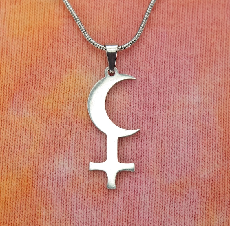 May include: A silver-colored pendant necklace featuring a crescent moon above a stylized cross. The pendant hangs from a silver chain. The background is a textured fabric with shades of pink, orange, and yellow.