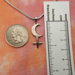 May include: A silver-colored pendant necklace featuring a crescent moon and cross symbol. The pendant is displayed next to a U.S. quarter and a ruler showing inches and centimeters. The necklace chain is a thin, silver snake chain.