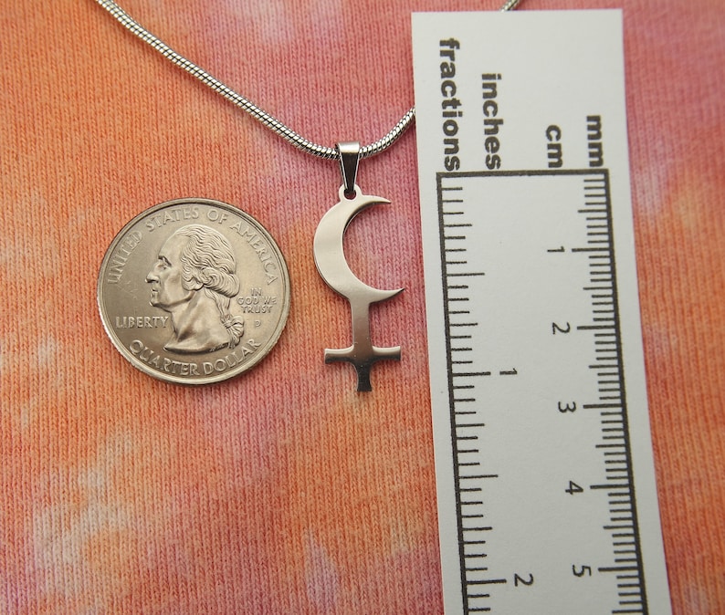 May include: A silver-colored pendant necklace featuring a crescent moon and cross symbol. The pendant is displayed next to a U.S. quarter and a ruler showing inches and centimeters. The necklace chain is a thin, silver snake chain.