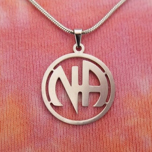 Könnte beinhalten: Eine silberne Halskette mit einem runden Anhänger, der die Buchstaben "NA" in einem stilisierten Design zeigt.