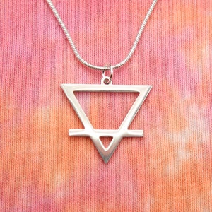 May include: A silver necklace with a geometric pendant in the shape of a triangle with a line across the middle. The pendant is on a silver chain and is hanging against a pink and orange tie-dye background.