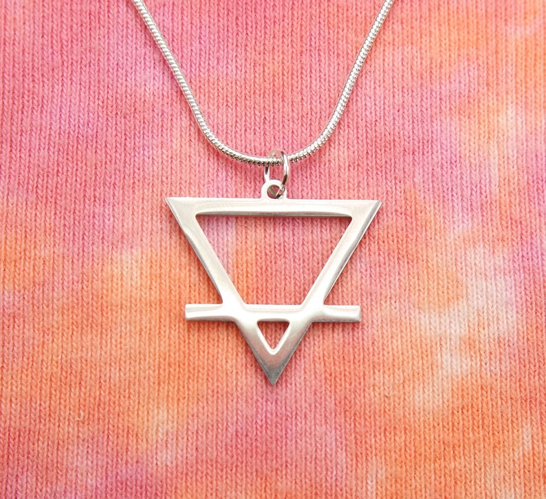 May include: A silver necklace with a geometric pendant in the shape of a triangle with a line across the middle. The pendant is on a silver chain and is hanging against a pink and orange tie-dye background.