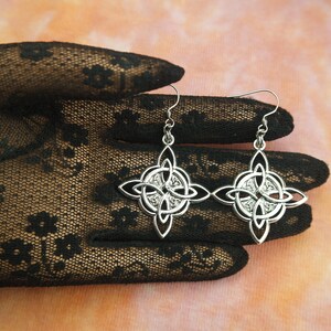 May include: A pair of silver Celtic knot earrings with a detailed design. The earrings are hanging from a black lace glove.