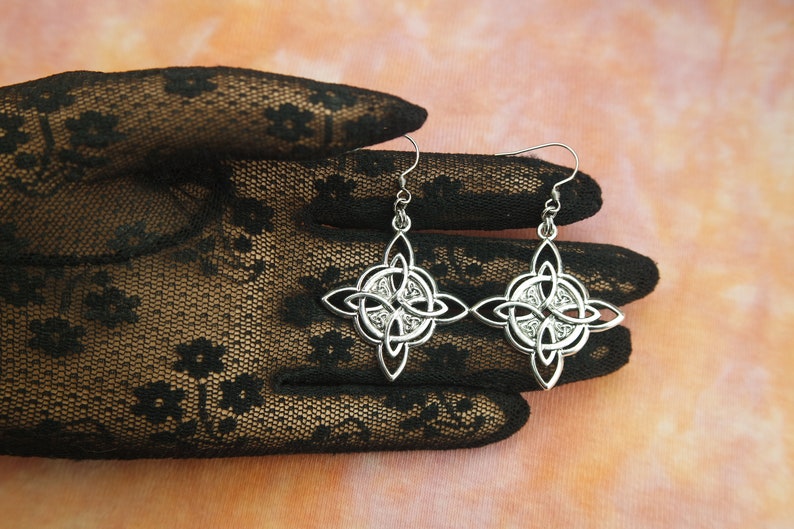 May include: A pair of silver Celtic knot earrings with a detailed design. The earrings are hanging from a black lace glove.