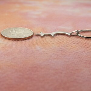 May include: A silver-colored pendant necklace with a unique, stylized key design. The pendant is next to a quarter for size comparison. The necklace chain is a thin, snake-style chain. The background is a soft, peach-colored fabric.