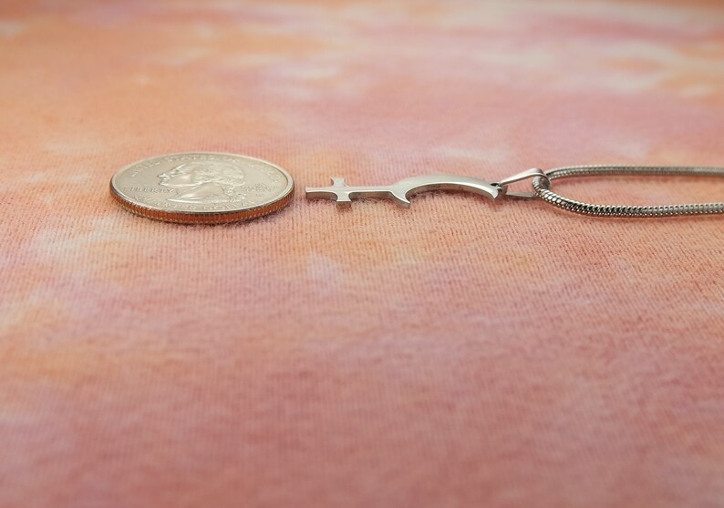 May include: A silver-colored pendant necklace with a unique, stylized key design. The pendant is next to a quarter for size comparison. The necklace chain is a thin, snake-style chain. The background is a soft, peach-colored fabric.