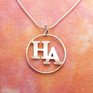 Może przedstawiać: Srebrny naszyjnik z okrągłym wisiorem, na którym wycięto litery "HA" w prostym kroju czcionki.