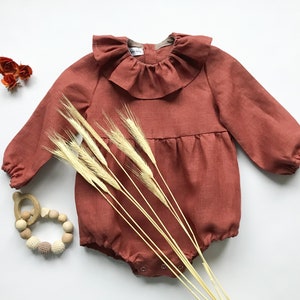 May include: Rust-colored baby romper with a ruffled collar and long sleeves. The romper has a gathered waist and snap closures. A wooden teething ring and wheat stalks are next to the romper.