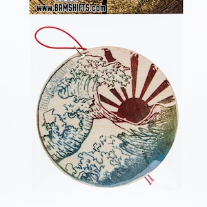 May include: A round wooden air freshener featuring a Japanese wave and rising sun design. The air freshener is beige with blue, red, and green accents. It has a red string attached for hanging.