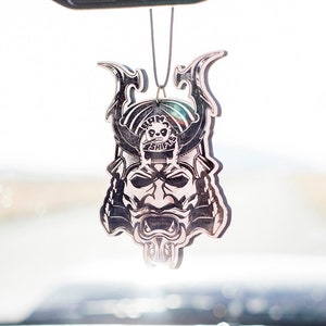 May include: A silver car air freshener with a detailed design of a samurai warrior's face. The design is intricate and includes a skull on the helmet.