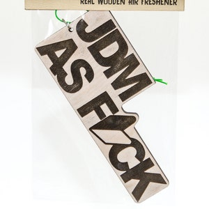 May include: A wooden car air freshener with the text "JDM AS F*CK" in black lettering.