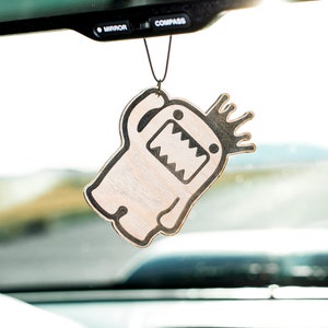 Puede incluir: Un ambientador de coche beige con un diseño de monstruo de dibujos animados en blanco y negro. El monstruo tiene una corona en la cabeza y está saludando.