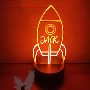 May include: A yellow rocket-shaped night light with the name "Jack" inscribed on it. The light is on a black base and is sitting on a red surface.