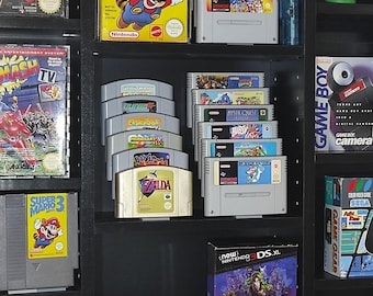 May include: A black shelf unit with various video game cartridges, including Super Mario 3, Zelda, and Mystic Quest. The shelf also features a Game Boy Camera and a Nintendo 3DS XL.