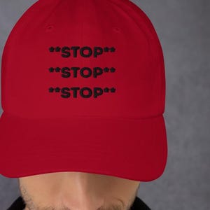 May include: A red baseball cap with the word "STOP" repeated three times in black, with asterisks on each side of the word. The cap has a curved brim and a fabric strap closure.