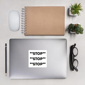 Könnte beinhalten: Ein weißer Aufkleber mit dem Wort "**STOP**" dreimal in fetter schwarzer Schrift wiederholt. Der Aufkleber befindet sich auf einem Laptop, umgeben von einer Computermaus, einem Stift, einem Notizbuch, einer Brille und kleinen Topfpflanzen. Die Gegenstände befinden sich auf einer hellgrauen Oberfläche.