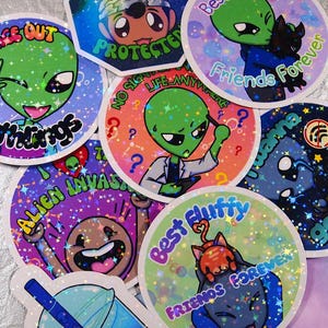 Op de afbeelding: Een verzameling kleurrijke, holografische stickers met cartoonaliens en andere grillige ontwerpen. De stickers bevatten zinnen als "Peace Out, Earthlings", "Best Fluffy Friends Forever" en "Alien Invasion". Een blauwe boba tea sticker is ook inbegrepen.