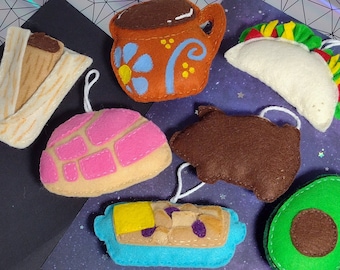 Op de afbeelding: Een set vilt ornamenten in de vorm van verschillende Mexicaanse voedingsmiddelen, waaronder een tamale, een concha, een kopje koffie, een taco, een avocado en een gebakje gevuld met chocolade. De ornamenten zijn allemaal met de hand gestikt en hebben een rustieke, handgemaakte uitstraling.
