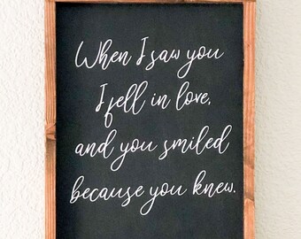 以下が含まれることがあります： ウィリアム・シェイクスピアの「When I saw you I fell in love and you smiled because you knew.」という引用句が書かれた木製の額縁入りサイン。サインは黒い背景に白い文字で書かれています。サインは、本や装飾品が置かれた棚の上に飾られています。