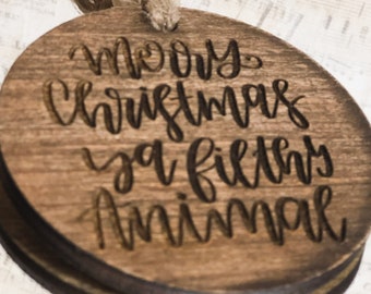 Op de afbeelding: Een ronde houten ornament met de tekst "Merry Christmas ya filthy animal" geschreven in zwarte cursieve letters.