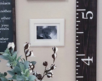 May include: A dark brown wooden growth chart with white numbers 3, 4, 5, and 6. The chart is mounted on a white wall with a framed picture of a child and a framed sign that says "it's so good to be home".