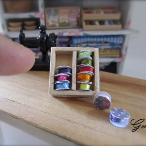 Peut inclure: Petite boîte de couture en bois avec des bobines de fil colorées. Les bobines sont dans les tons de violet, rose, rouge, orange, vert et jaune. Deux bobines décoratives sont à côté de la boîte. Une machine à coudre noire miniature est en arrière-plan.