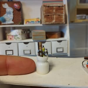 Puede incluir: Un tarro de cerámica blanco con tapa plateada contiene una colección de lápices y una regla. El tarro está sobre una superficie blanca.