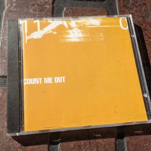 Op de afbeelding: Een gele CD-doos met de tekst "COUNT ME OUT" erop gedrukt.